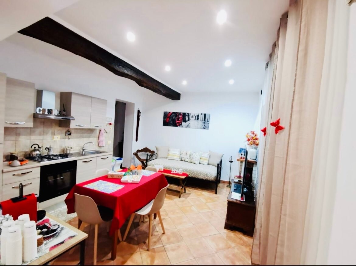 The open living area features a modern kitchen with essential appliances and a dining table set for meals. A comfortable sofa enhances the space, accompanied by soft lighting from ceiling fixtures. Warm tones and wooden beams contribute to a welcoming environment.