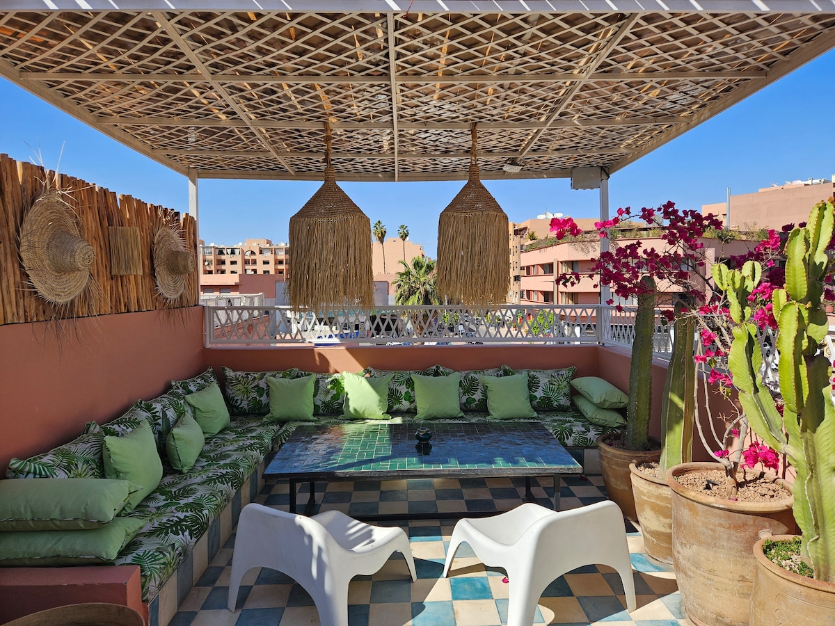 A rooftop terrace is displayed, featuring a low seating area surrounded by lush green cushions. Decorative straw elements hang above, while vibrant bougainvillea and a cactus add natural touches. The space is illuminated by sunlight, showcasing a colorful tiled floor.