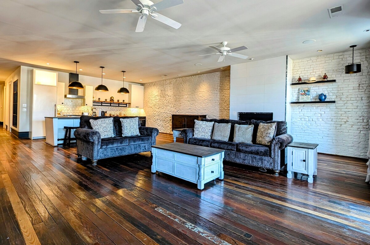 An inviting living area features plush sofas adorned with decorative pillows. A wooden coffee table sits center stage on rich hardwood flooring, complemented by exposed brick walls. In the background, the gourmet kitchen is visible, illuminated by pendant lighting.