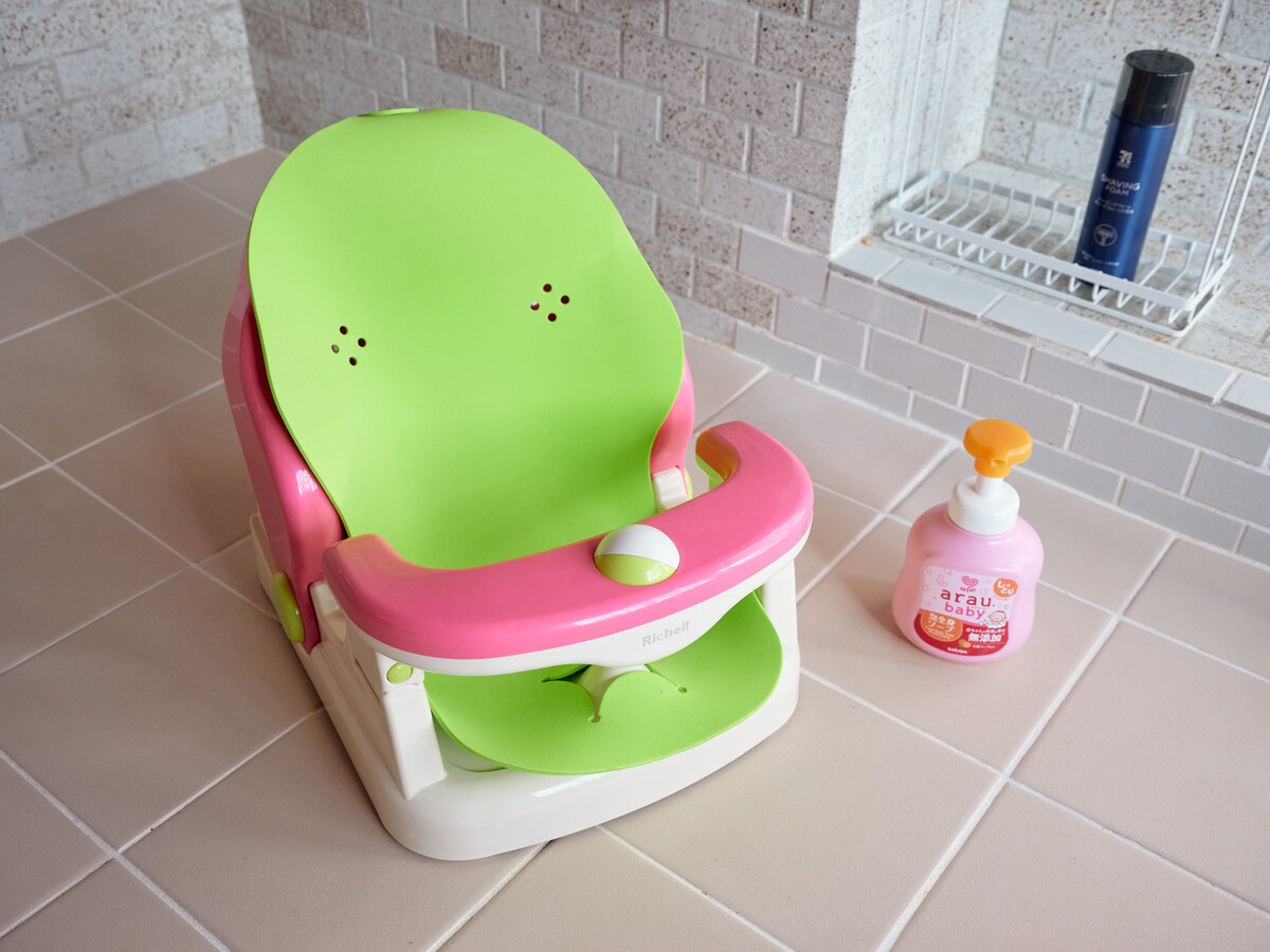 A colorful baby high chair features a green and pink design, set on tiled flooring. Next to it, a bottle of hand soap sits on the counter, providing a convenient place for hygiene. The clean, modern bathroom space is enhanced by subtle tile textures.