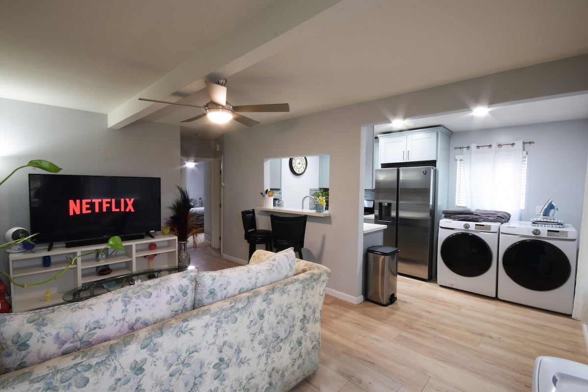 A spacious living area is equipped with a 65-inch Smart TV displaying the Netflix logo. A floral-patterned sofa sits comfortably in front of the TV. An open kitchen area is visible in the background, featuring stainless steel appliances, including a full-size fridge and a washer/dryer.