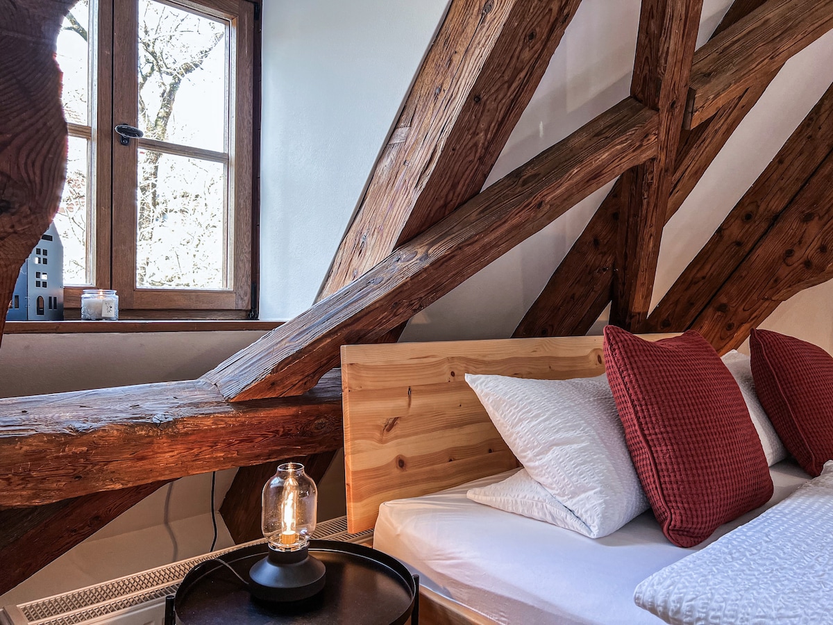 A sleeping area features exposed wooden beams that highlight the room's architecture. A spacious bed is adorned with red checkered pillows, and a small round table with a lantern rests beside it. Natural light enters through the nearby window, providing a calming ambiance.