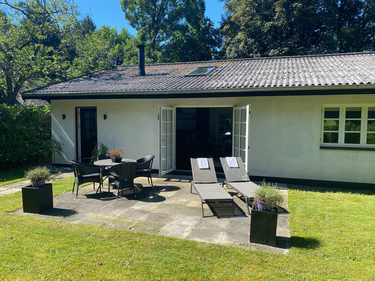 A patio area is presented with two sunbeds and a round dining table surrounded by chairs. A well-maintained lawn and flowering plants are visible. The villa features double doors leading to the interior, providing an inviting transition to the outdoor space.