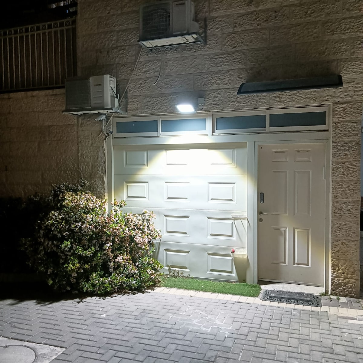 A small entrance is visible, featuring a light-colored door with a single handle and a nearby window above. A garage door is positioned adjacent, illuminated by an overhead light. Lush greenery grows near the entrance, complementing the outdoor stone pathway.