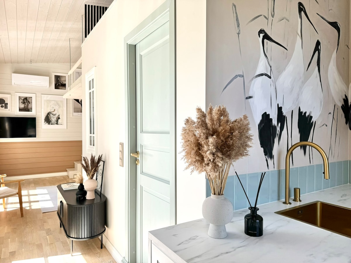 A bright and airy interior is highlighted by soft pastel colors and a stylish kitchenette. Decorative elements, including a vase of dried flowers and a black and white artwork featuring cranes, are visible, adding an artistic touch. A subtle division between the living area and kitchen is suggested by the layout.