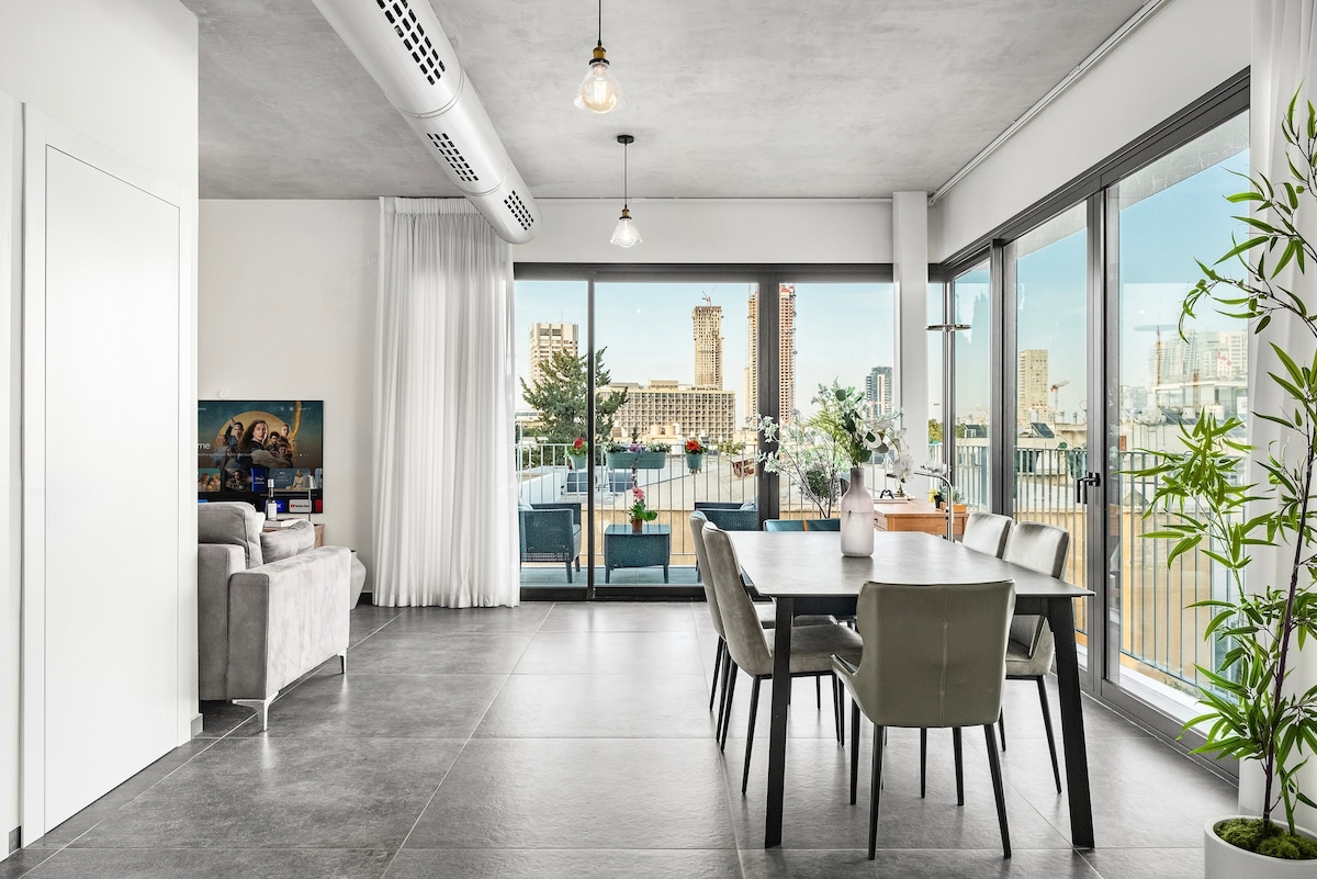 A spacious living and dining area features large glass windows allowing natural light to illuminate the room. A modern dining table with four chairs is positioned near the windows, with a comfortable seating arrangement visible in the adjacent space. Outdoor seating is seen beyond the glass.