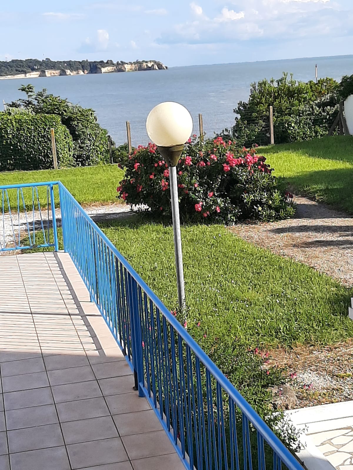 A view of the garden area includes well-maintained greenery and blooming flowers, with a pathway leading towards the sea. A lamp post stands beside the pathway, providing subtle illumination. In the distance, the coastal cliffs are visible against the serene waters of the sea.