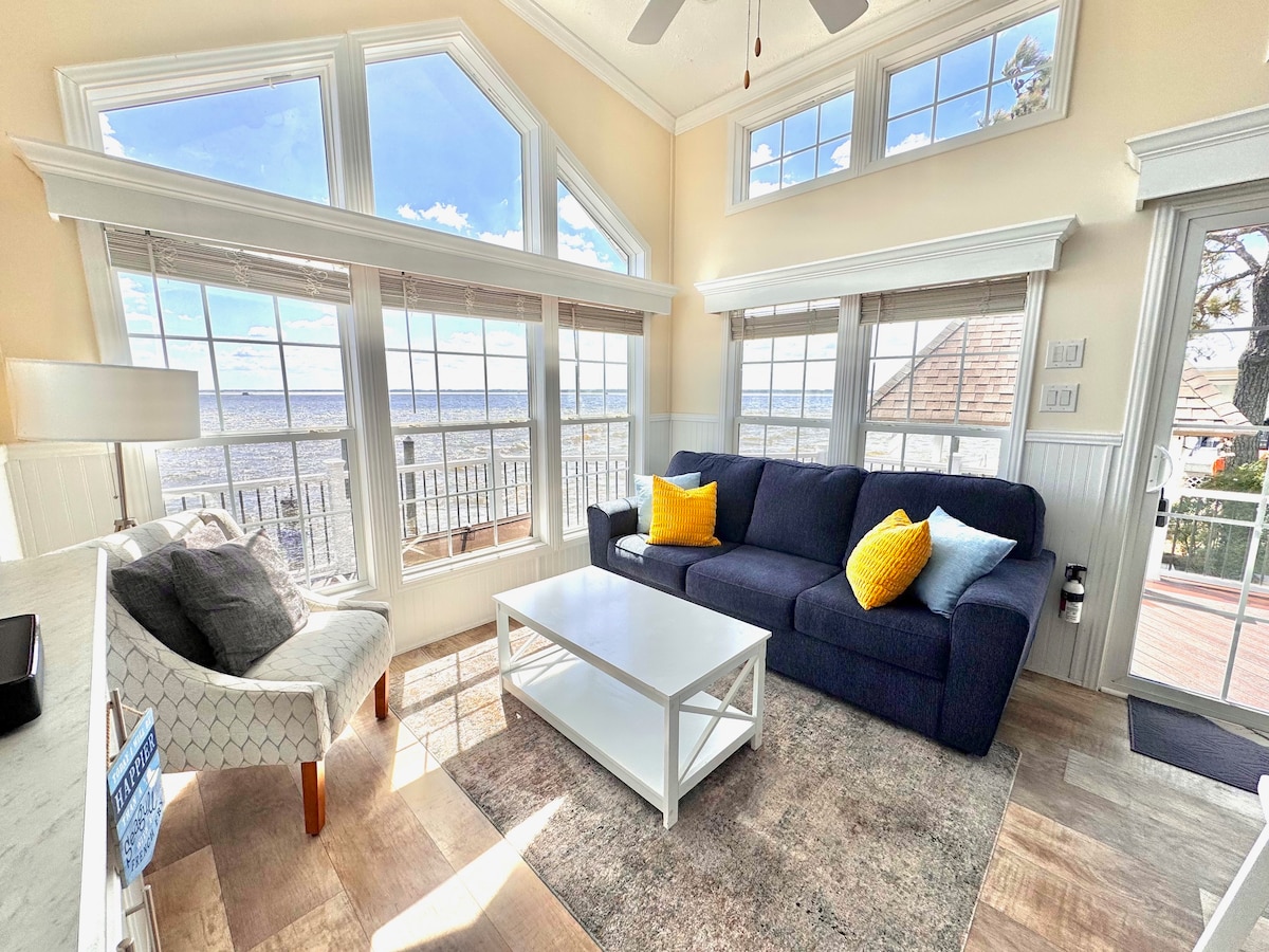A bright living area features large windows providing expansive views of the bay. A dark blue sofa with colorful accent pillows is positioned beside a white coffee table over a soft area rug. Light floods in, enhancing the open and airy atmosphere.