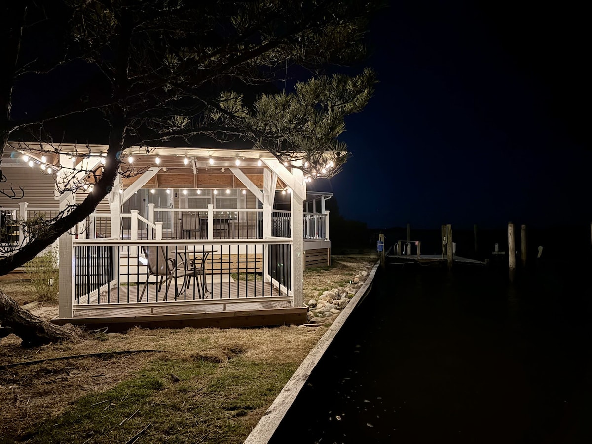A wooden deck extends from the tiny home, illuminated with string lights. The area features a table and chairs, suitable for outdoor dining. A path leads to the water's edge, where wooden posts and a rippling surface create a serene nighttime ambiance.