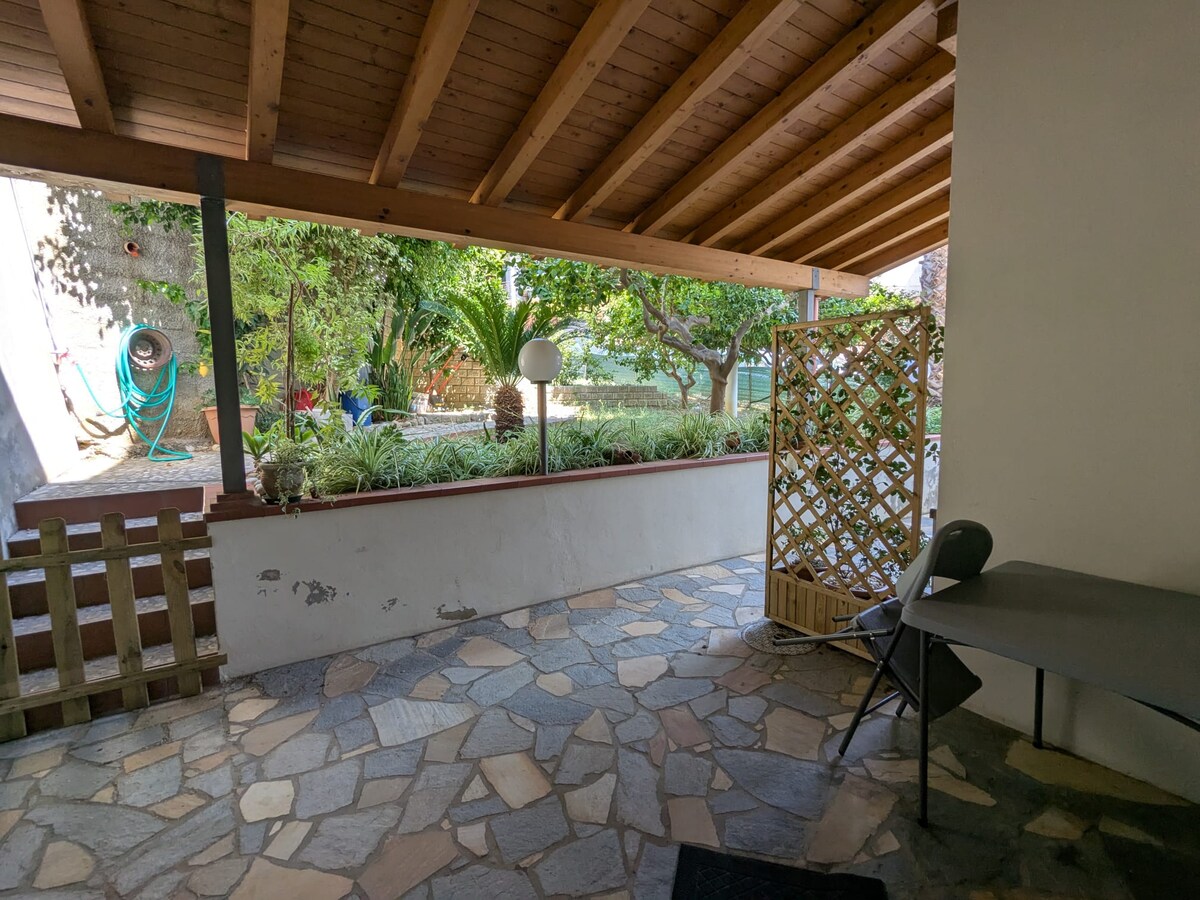 An outdoor patio area features a stone floor and a wooden ceiling. A decorative lattice provides partial privacy, while a small table and chair are positioned against the wall. Lush greenery and palm trees can be seen beyond the patio, enhancing the tranquil setting.