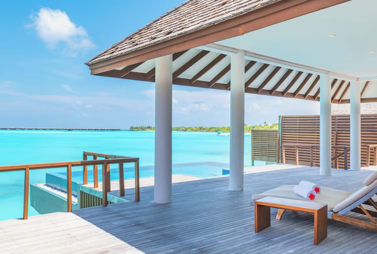 A spacious outdoor deck is shown, featuring wooden flooring and shaded areas. A sun lounger is positioned nearby, adorned with rolled towels. The vibrant blue water stretches into the distance, framed by lush greenery and a clear sky, enhancing the serene environment.