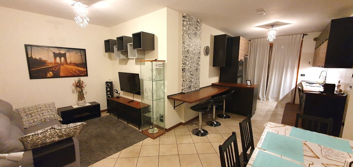 An elegantly furnished living area features a gray sofa, a glass display cabinet, and a black television unit. A compact workspace with a black chair is situated beside the kitchen area. Light tiles cover the floor, while soft overhead lighting creates a warm environment.