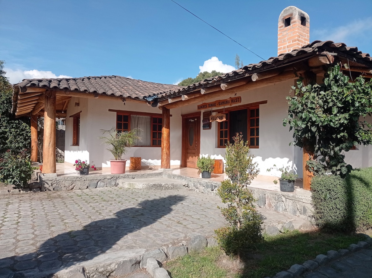 A rustic house is showcased with a tiled roof and wooden beams. The entrance features a welcoming door, flanked by potted plants and greenery. A stone pathway leads to the home, surrounded by landscaped areas, offering a tranquil atmosphere.