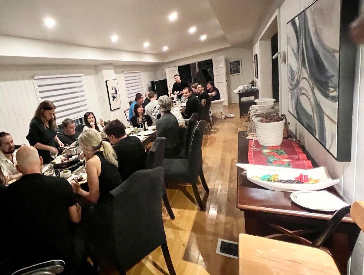 A long dining table is filled with guests enjoying a meal in a spacious, well-lit room. Dark upholstered chairs surround the table, while large windows with white shades allow for natural light. A buffet table with refreshments is set against a wall decorated with artwork.