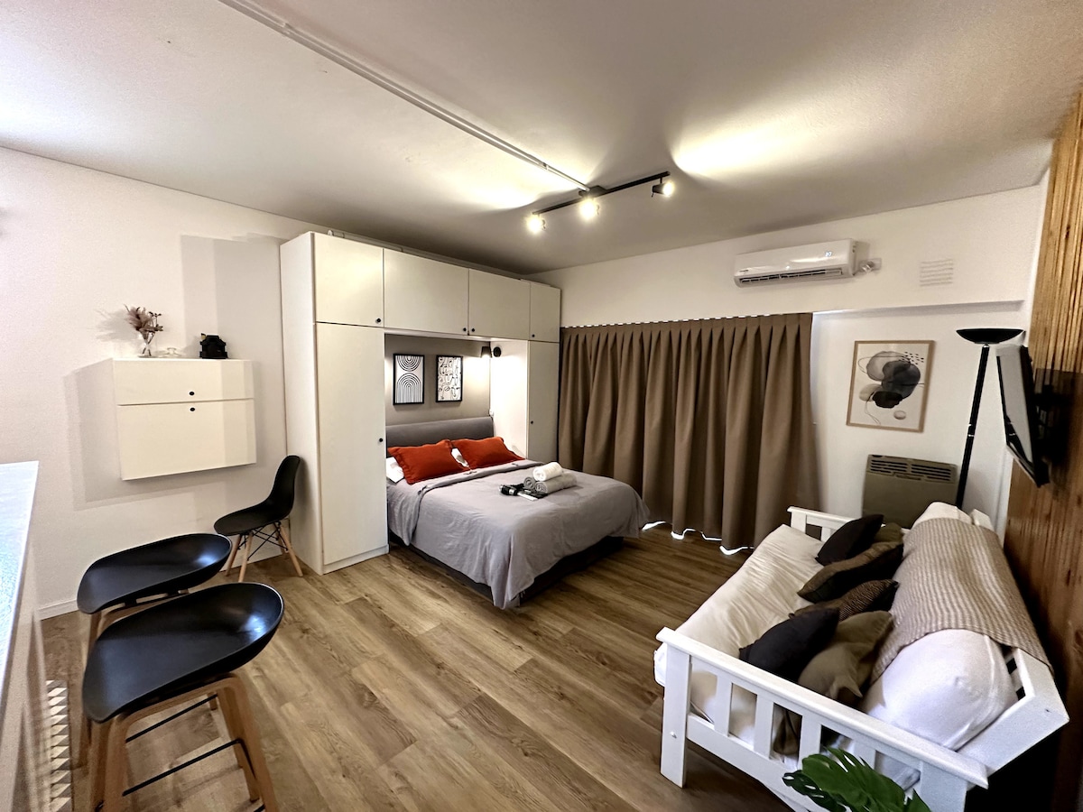 The studio features a spacious bed with neutral bedding and contrasting decorative pillows. A futon sofa is positioned beside it, offering additional sleeping space. A compact dining area with two chairs is visible, along with wooden flooring and natural light filtering through the curtains.