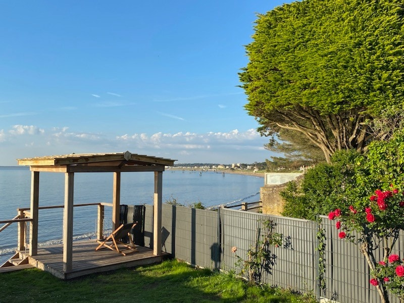 A wooden gazebo is positioned by the water's edge, featuring two chairs for relaxation. Lush greenery surrounds the area, with blooming roses adding color. The tranquil sea extends into the distance, offering scenic views under a clear blue sky.