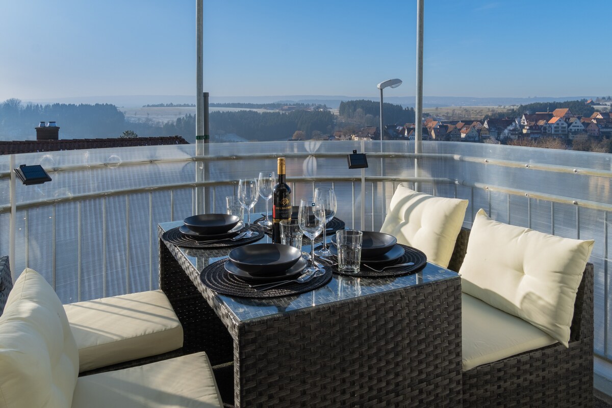 An outdoor balcony is furnished with a glass-topped dining table set for four, featuring dinnerware and a bottle of wine. Comfortable cushioned seating is arranged around the table, offering expansive views of the landscape and distant hills under a clear blue sky.