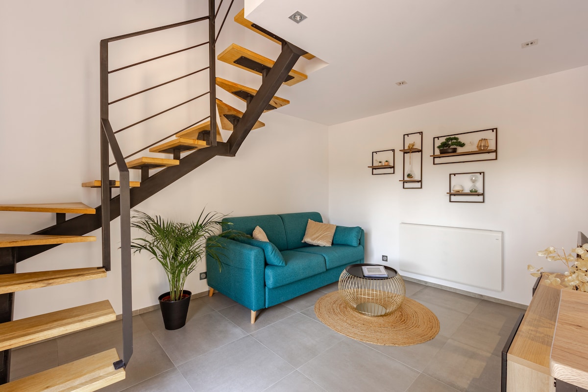 A modern living area features a light-colored wall and a stylish staircase with wooden treads and a metal railing. A teal sofa is positioned next to a round glass table on a woven rug. Shelves displaying decorative items are mounted on the wall.