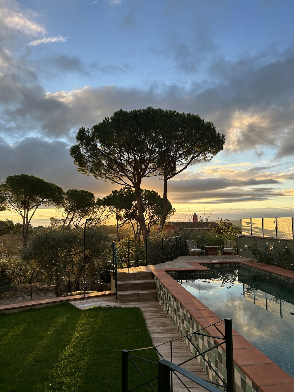 A serene outdoor space features a well-maintained garden and a swimming pool reflecting the sky. A large tree is prominently positioned alongside the pool, with expansive views of the surrounding landscape. The scene is illuminated by soft evening light.