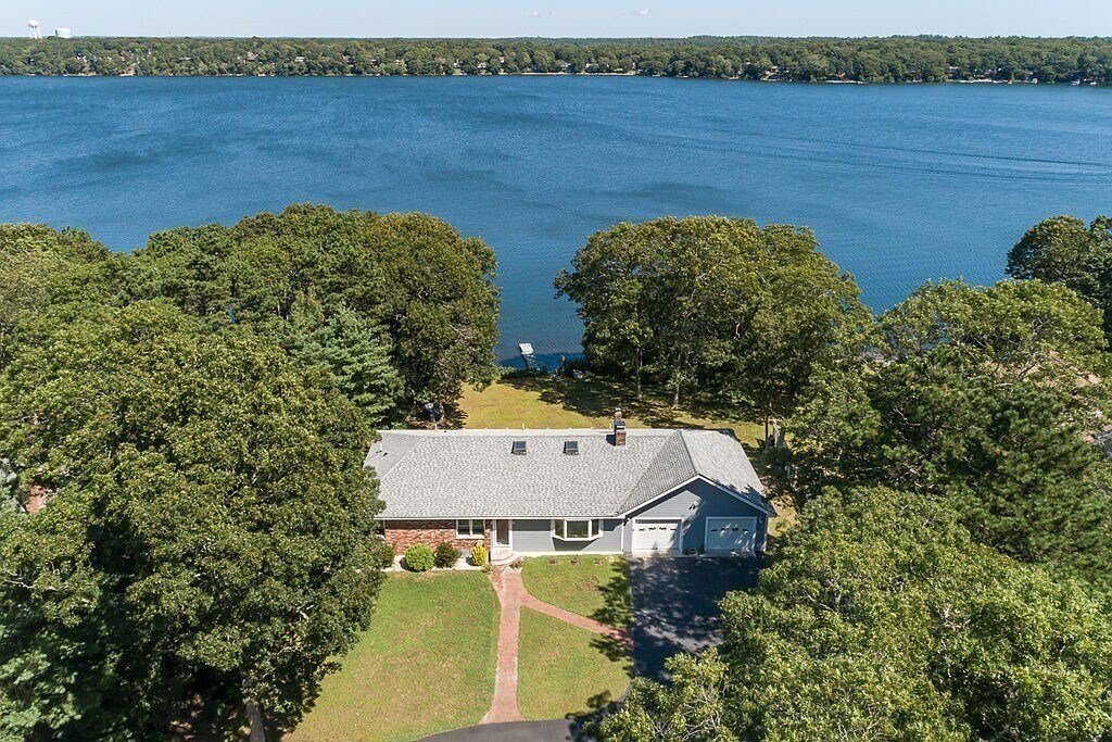 An aerial view showcases a single-story home surrounded by lush greenery, with the tranquil lake visible in the background. The neatly manicured lawn leads up to the house, which features a driveway and a garage. The expansive water surface reflects the bright sky.