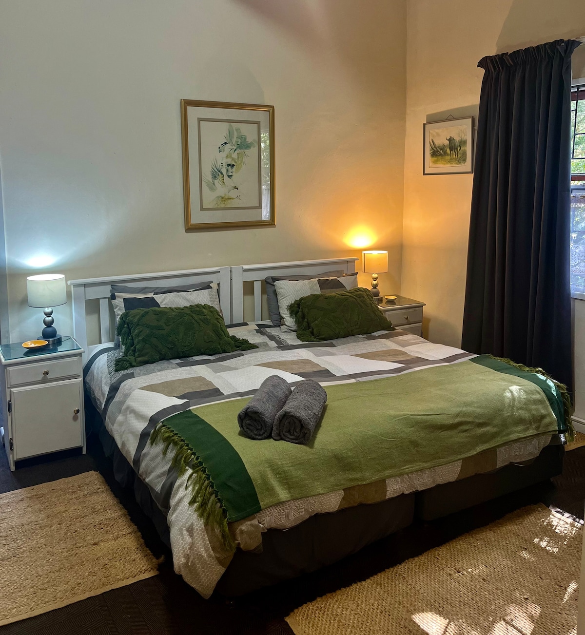 A cozy sleeping area is presented with a spacious bed dressed in a patchwork blanket of neutral and green tones. Two bedside lamps provide soft lighting, while framed artwork decorates the walls. Towels are neatly arranged on the bed, and dark curtains frame the window.