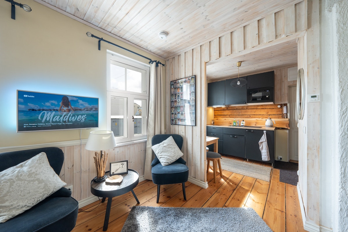 The living area features wooden paneling and warm lighting, with two armchairs beside a small table. A smart TV is mounted on the wall, and the adjoining kitchen is visible, showcasing modern cabinetry and wood accents.