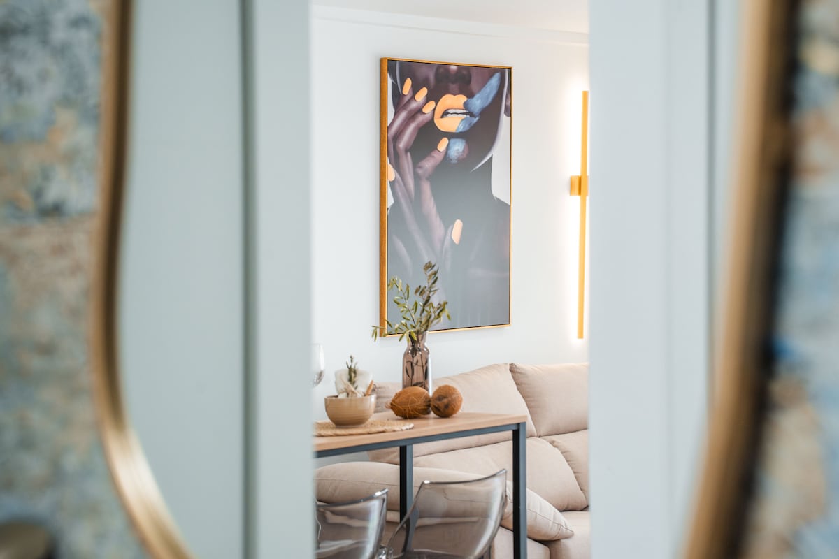 A modern living area is separated by a doorway, showcasing a sleek dining table with transparent chairs. A large piece of artwork featuring abstract hands hangs on the wall, while a cozy sofa with neutral tones is partially visible, contributing to a contemporary aesthetic.