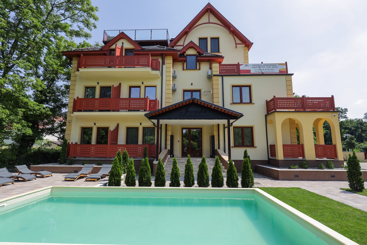 The villa is showcased with a well-maintained facade featuring multiple balconies adorned with red railings. A clear swimming pool is situated in the foreground, surrounded by neatly trimmed hedges and lounge chairs. Lush trees provide a natural backdrop to the elegant structure.