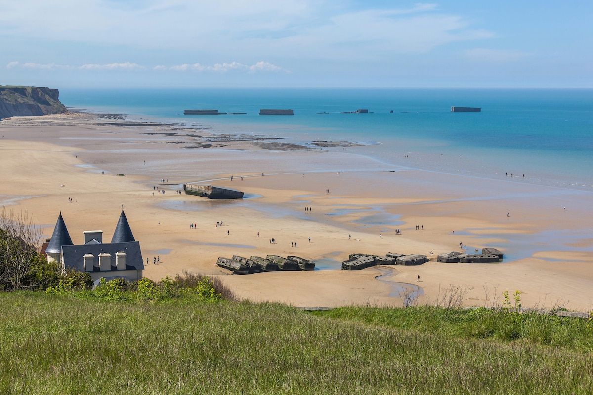 A serene beach scene is presented, featuring gentle waves lapping at a sandy shore. Historical structures are seen lying partially submerged in the water, with distant cliffs framing the coastline. A clear blue sky overhead enhances the tranquil atmosphere, inviting exploration and relaxation.