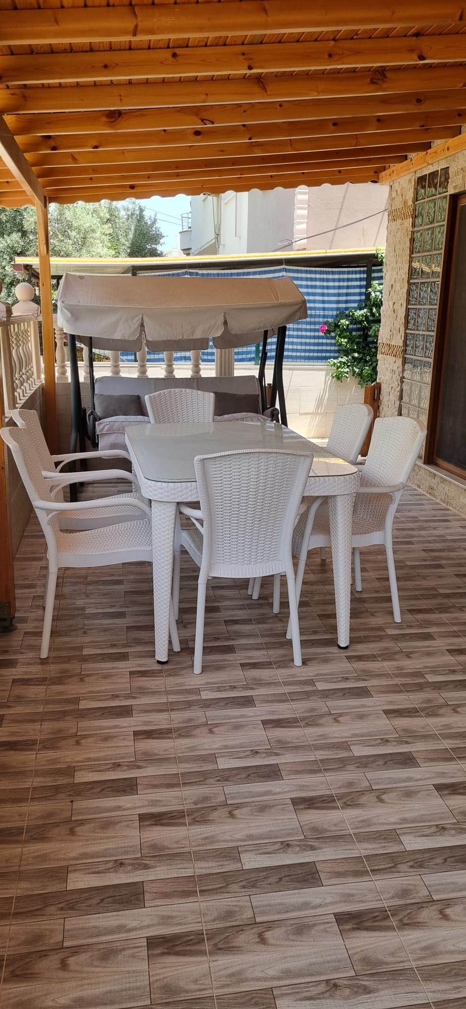 An outdoor space features a glass-top table surrounded by six white chairs. In the background, a swing seat is positioned under a wooden ceiling. The stone floor is complemented by a swimming pool and greenery visible beyond the patio.