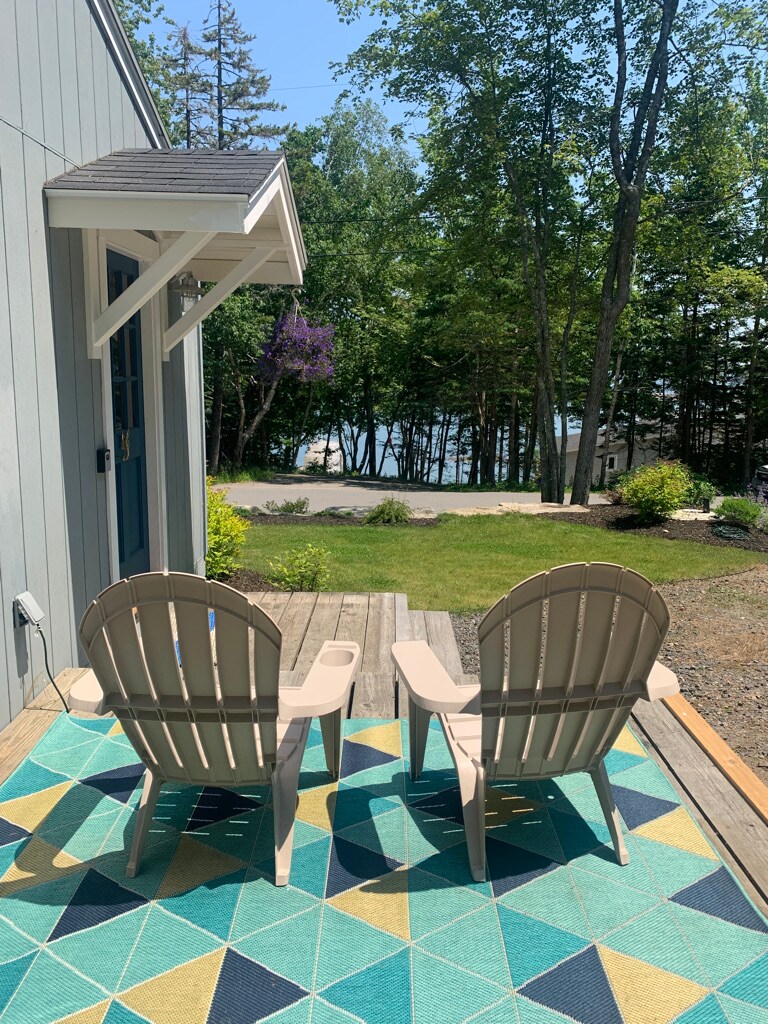 Two beige adirondack chairs are positioned on a patterned outdoor rug on a wooden deck. In the background, a view of a grassy area leads to a pathway and the shimmering water. Surrounding trees provide a natural, serene backdrop.
