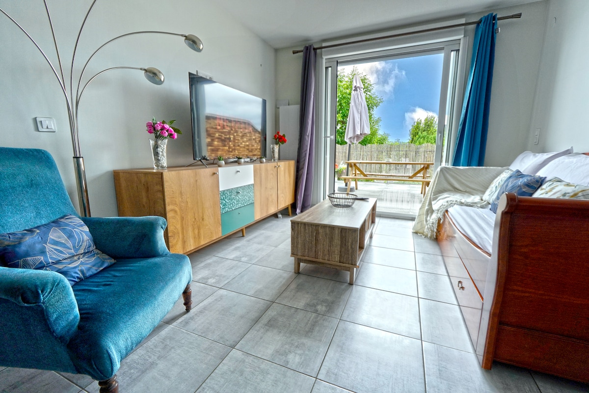 The living area features a blue armchair beside a wooden console table with flowers. A large TV is mounted on the wall, facing a convertible sofa. A coffee table is centered on the tile floor, while sliding glass doors open to a balcony with outdoor seating.