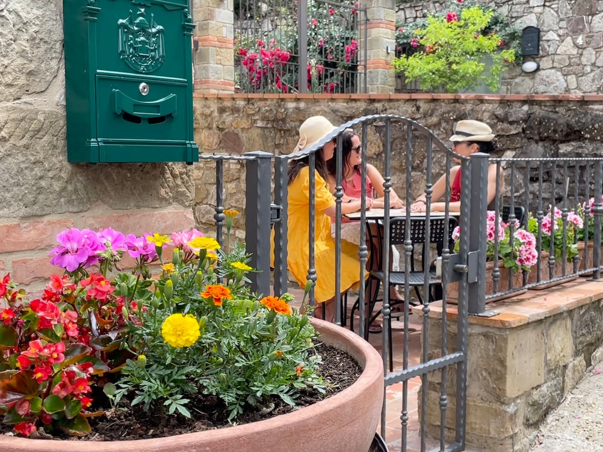 A charming outdoor seating area is framed by a decorative iron gate. Vibrant flowers in terracotta pots add a burst of color, while two individuals enjoy a moment together at a small table. A green mailbox is mounted on the stone wall, enhancing the inviting garden setting.