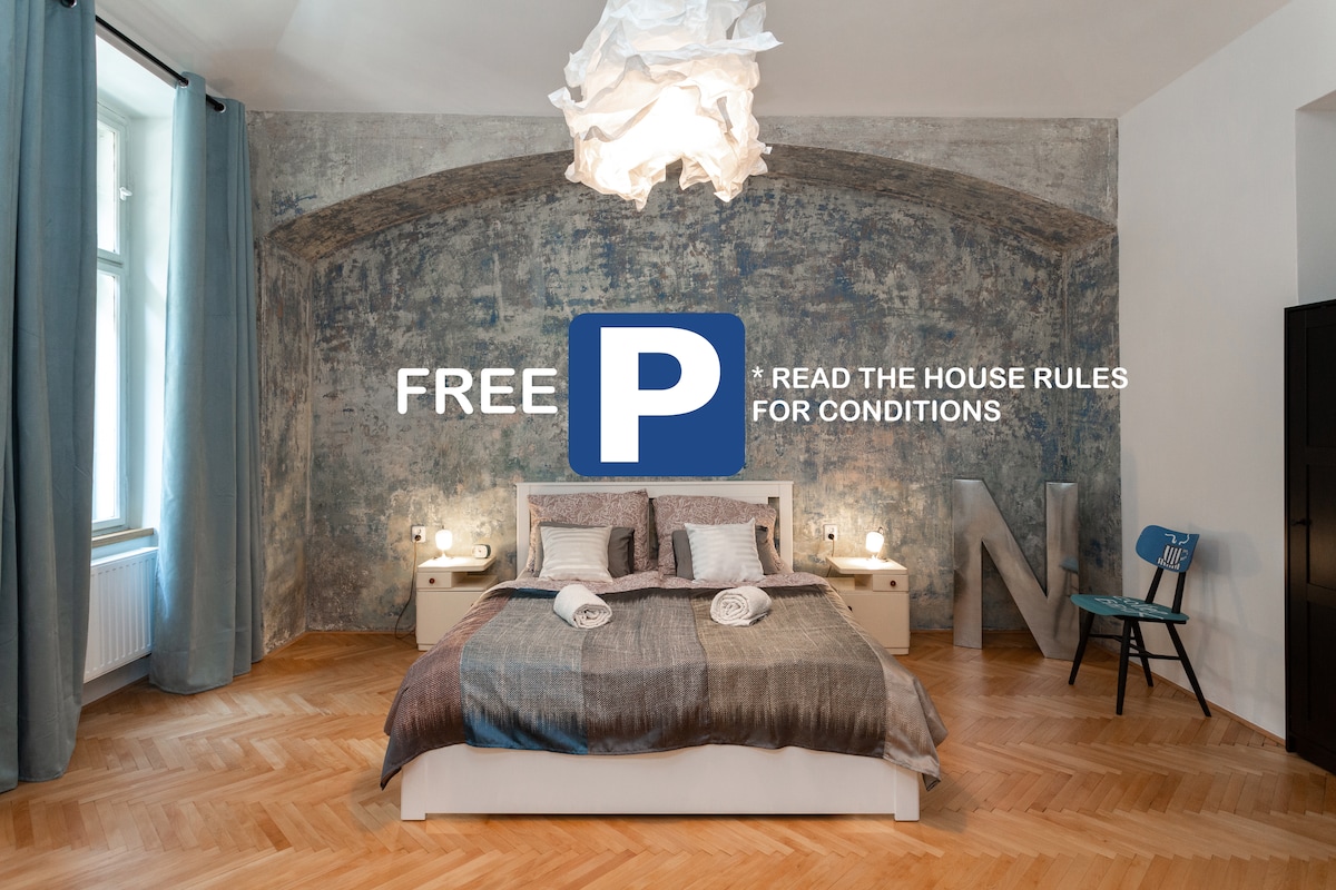 A cozy sleeping area features a spacious bed adorned with a striped blanket. Soft lighting enhances the textured blue wall and high arched ceiling. A playful accent chair and large letter add artistic flair, while natural light enters through the nearby window draped with light curtains.