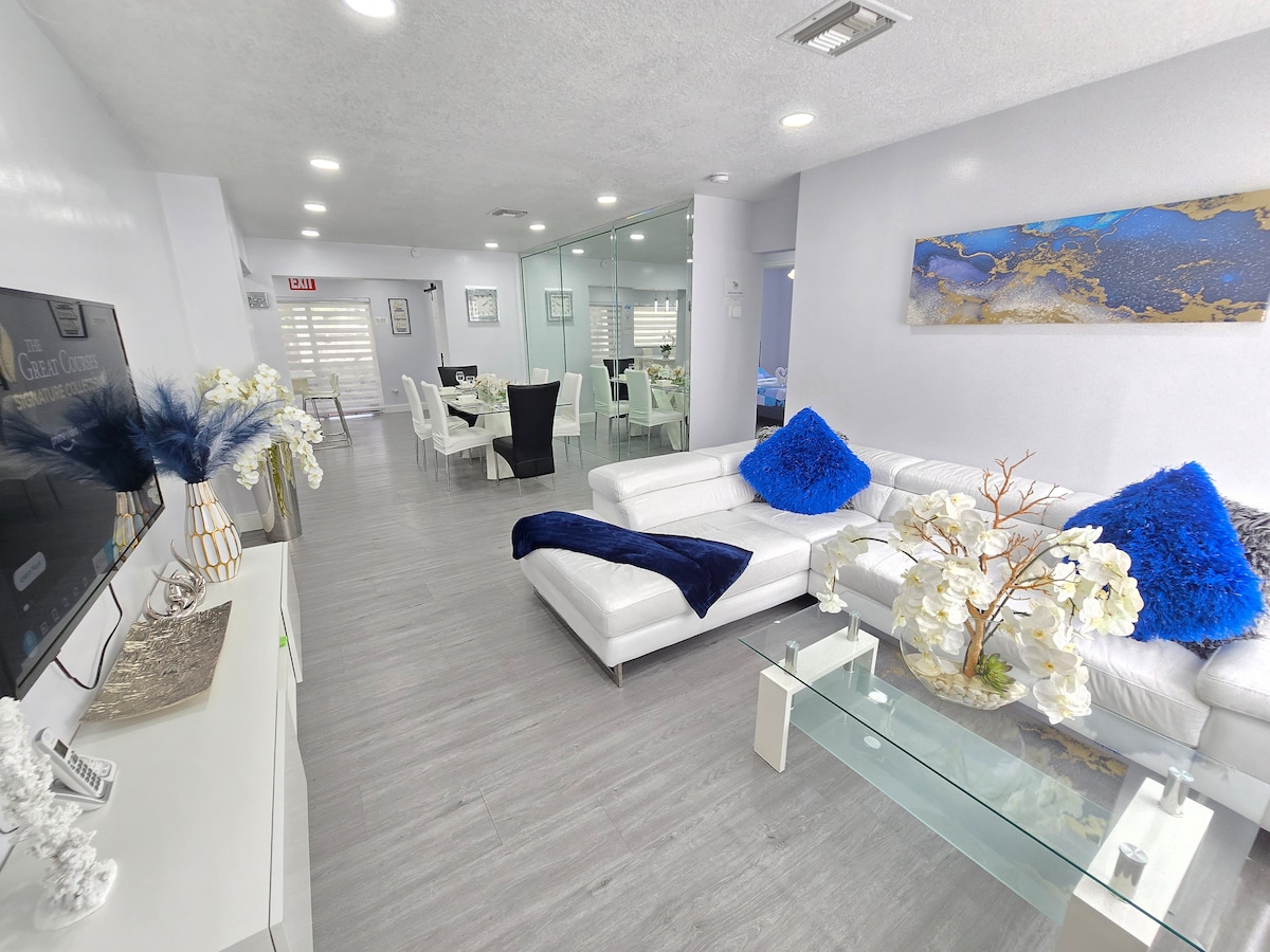A spacious living area is featured, showcasing a white sectional sofa adorned with blue accent pillows. A glass coffee table sits atop a light grey floor, complemented by decorative white flowers. A dining area is visible in the background, featuring a dark table set for six.