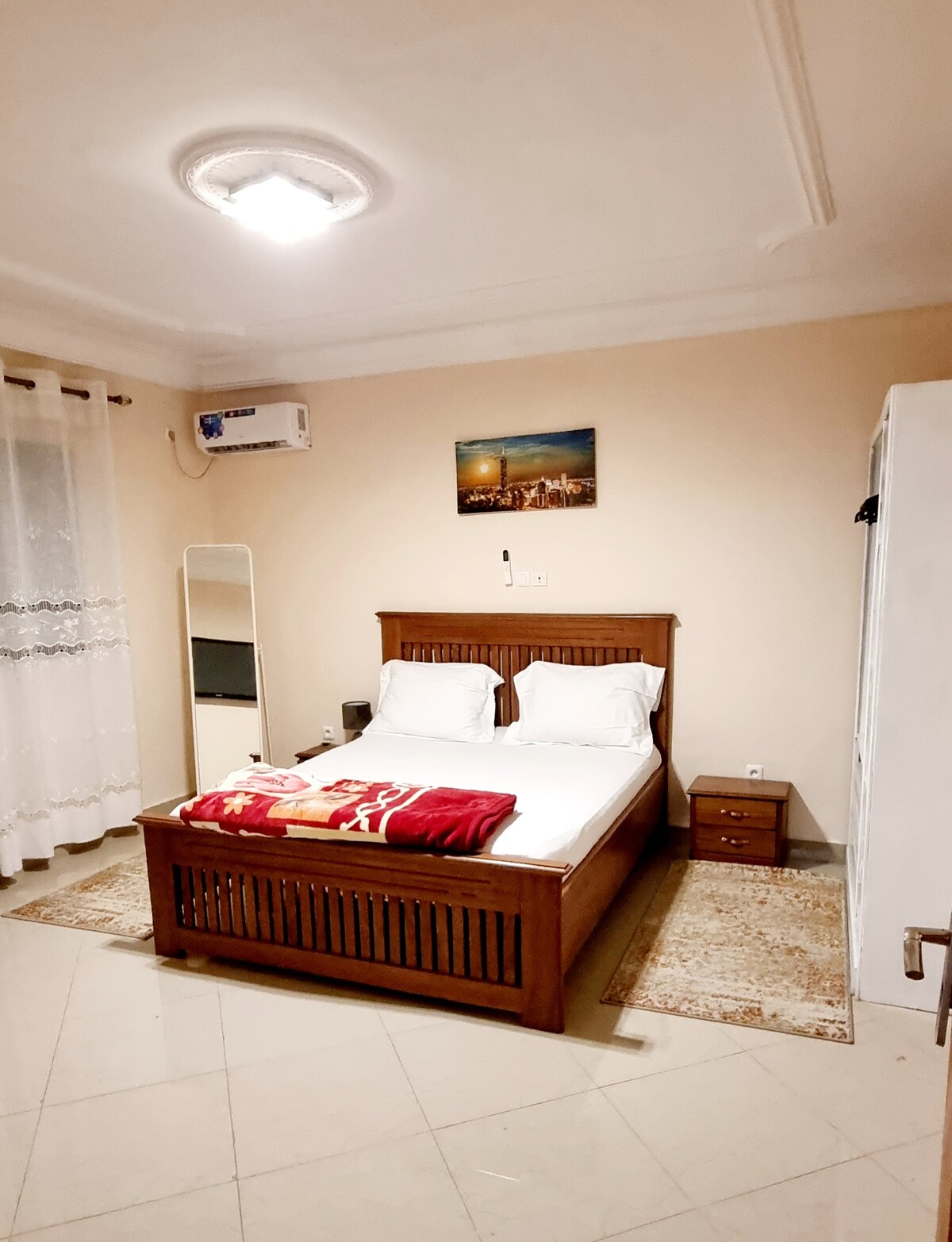 A spacious bedroom features a wooden bed with white bedding. A simple bedside table and a tall mirror are present. Natural light enters through sheer curtains, and a wall painting adds a touch of color to the room's neutral tones. An air conditioning unit is visible.