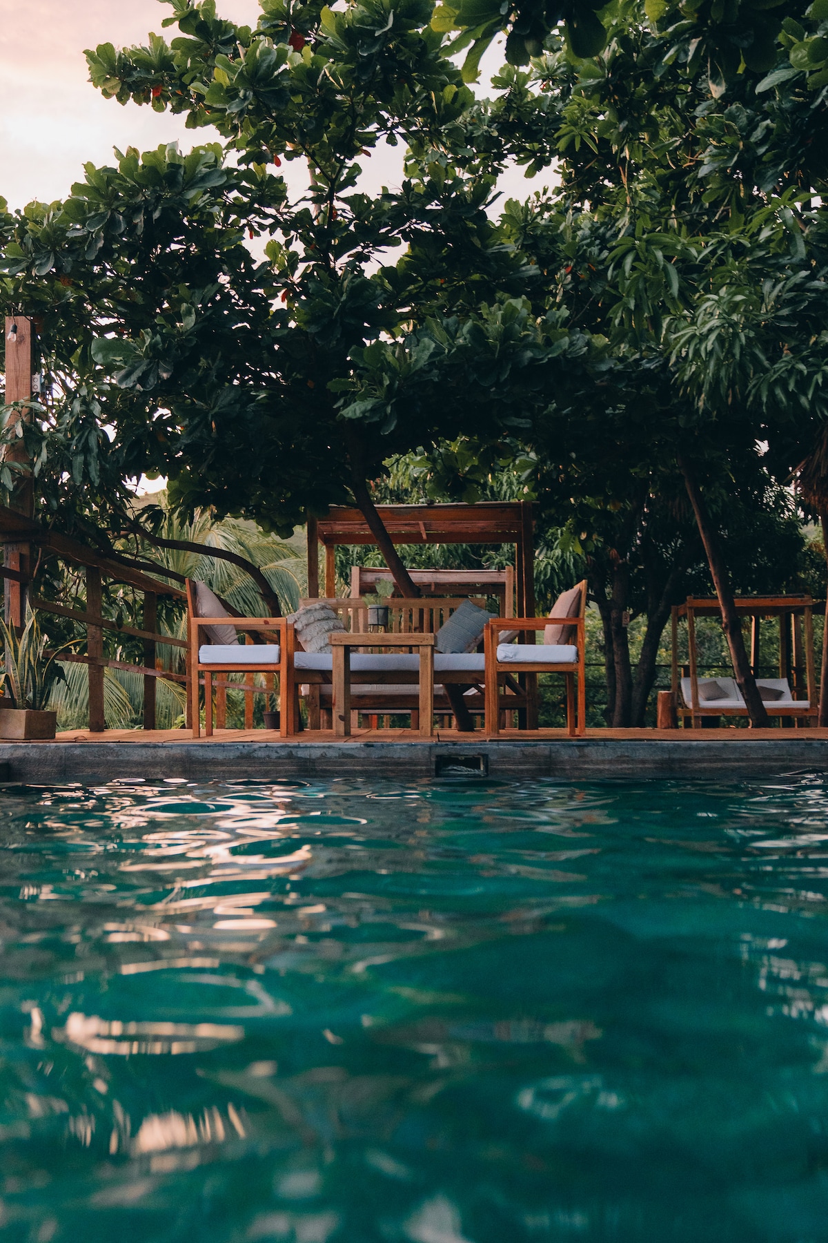 The swimming pool is surrounded by lush greenery, featuring a shaded seating area with wooden furniture. The water reflects the natural light, creating a serene atmosphere. The arrangement includes a table and chairs, ideal for relaxation beside the pool.