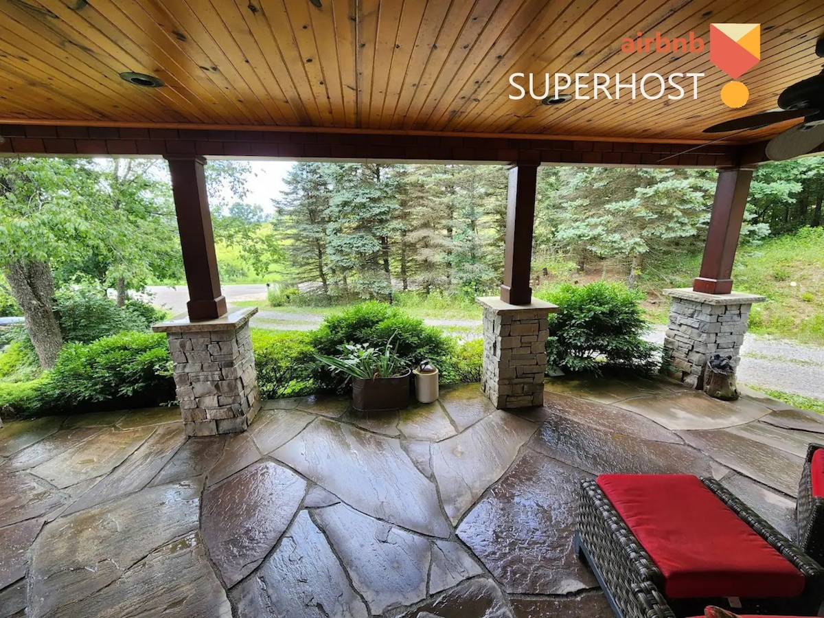 A spacious front patio is surrounded by lush greenery and stone columns. The natural stone flooring is complemented by a comfortable chair, providing an inviting place to relax and enjoy the scenic view of the landscape beyond.