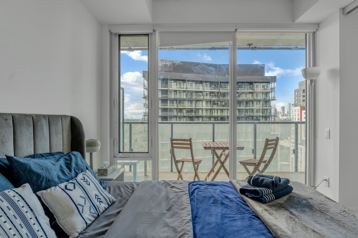 A cozy bedroom is enhanced by a large window that opens to a private balcony. Soft bedding in shades of blue and gray adorns the spacious bed. A small table and chairs on the balcony provide a spot for outdoor relaxation, surrounded by city views.