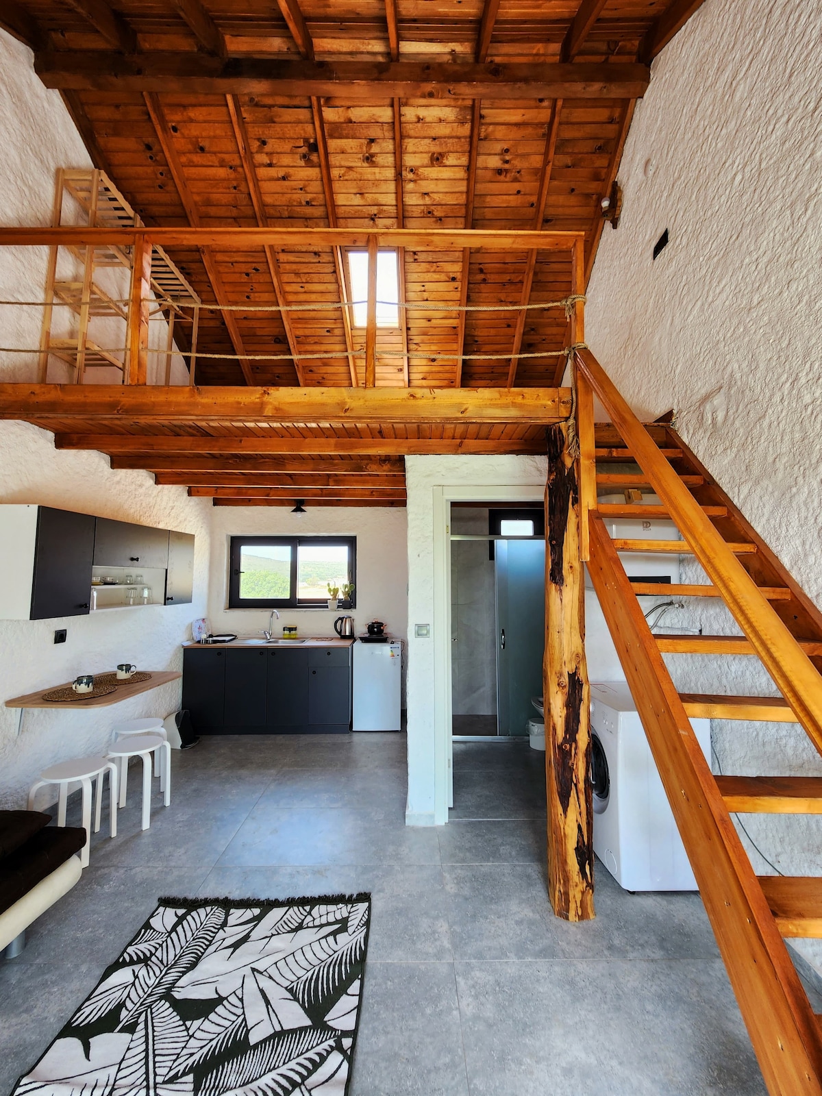 An open living area features high wooden ceilings and a staircase leading to a loft space. The kitchen area includes modern appliances, while a small dining table accommodates several chairs. A patterned rug is situated on the light gray flooring, enhancing the cozy atmosphere.