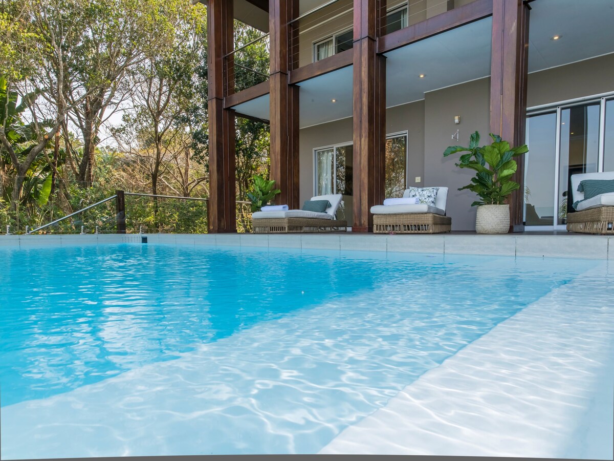 A tranquil private pool glistens under natural light, inviting relaxation. The surrounding area features comfortable outdoor seating with plush cushions, framed by lush greenery. The villa's modern architectural elements are visible, creating a harmonious blend with the serene environment.