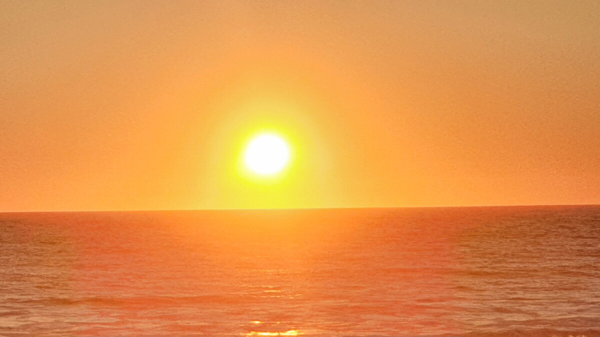A vibrant sunset casts a warm golden glow over the horizon, illuminating the calm ocean waters. The sun, prominent and round, creates a serene atmosphere as daylight transitions into evening, highlighting the peaceful beauty of the seaside.