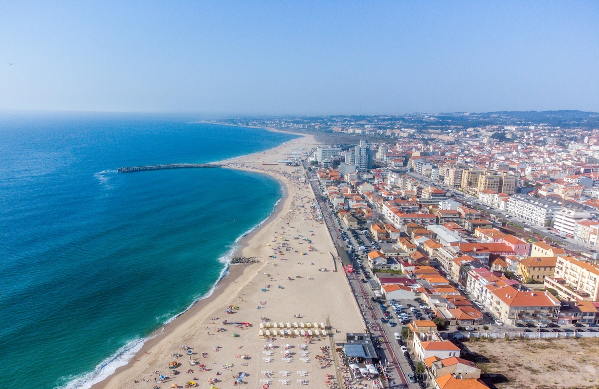 An aerial view captures the coastline, showcasing a sandy beach bordered by the sea. Colorful beach umbrellas dot the shore, while a series of buildings line the beachfront. The scene reflects a lively atmosphere with sunbathers, and the horizon extends to meet the clear blue sky.