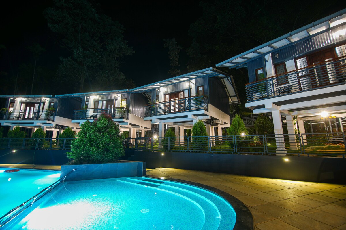 A row of modern accommodations is illuminated at night, each featuring balconies and large windows. A circular pool in the foreground reflects the ambient light, while landscaped greenery enhances the serene atmosphere of the property.
