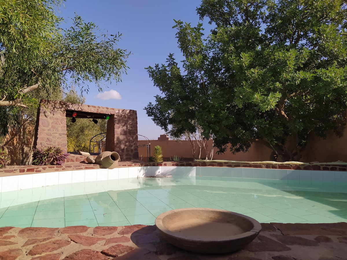 A serene outdoor area features a clear pool framed by natural stone edges. Lush greenery surrounds the space, with a stone archway visible in the background. A simple earthenware bowl is placed at the pool's edge, adding to the tranquil ambiance.