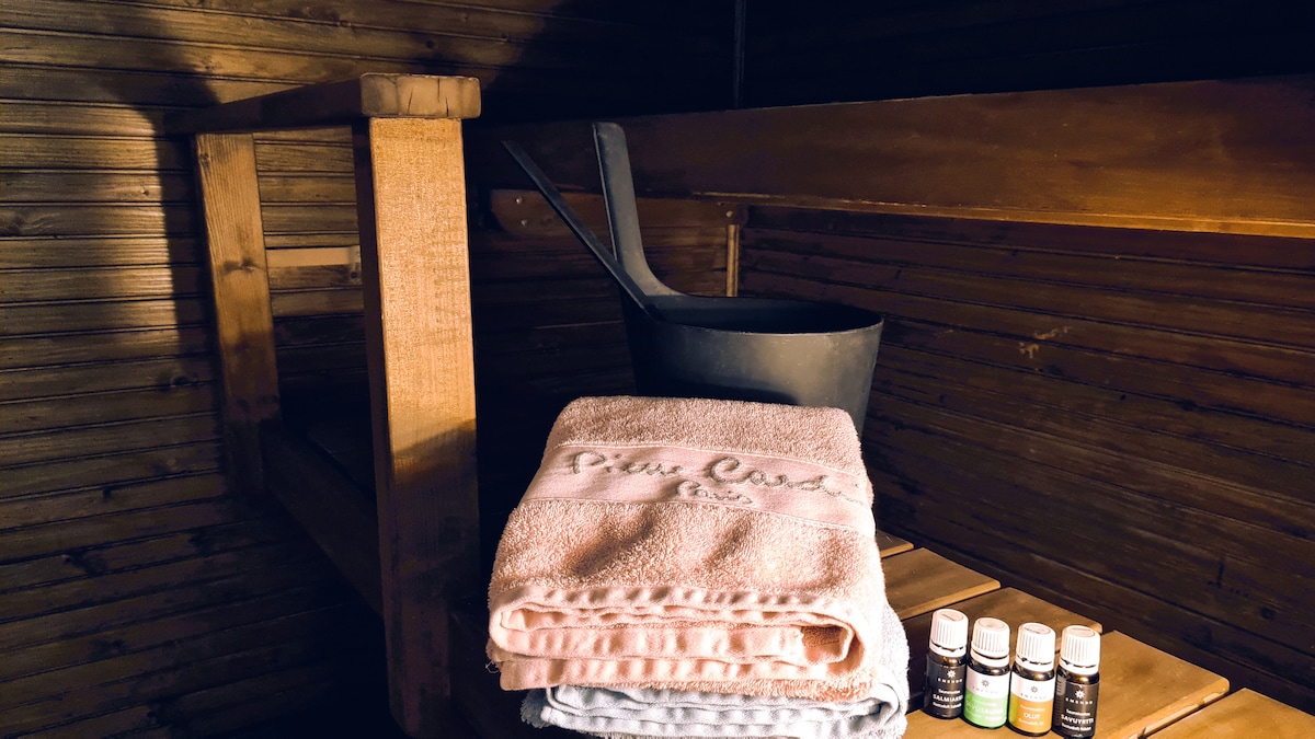 A wooden sauna interior is depicted, featuring a wooden bench and a metal bucket. Soft towels in light pastel colors are stacked on the bench, alongside small bottles of essential oils, adding to the overall ambiance of relaxation.