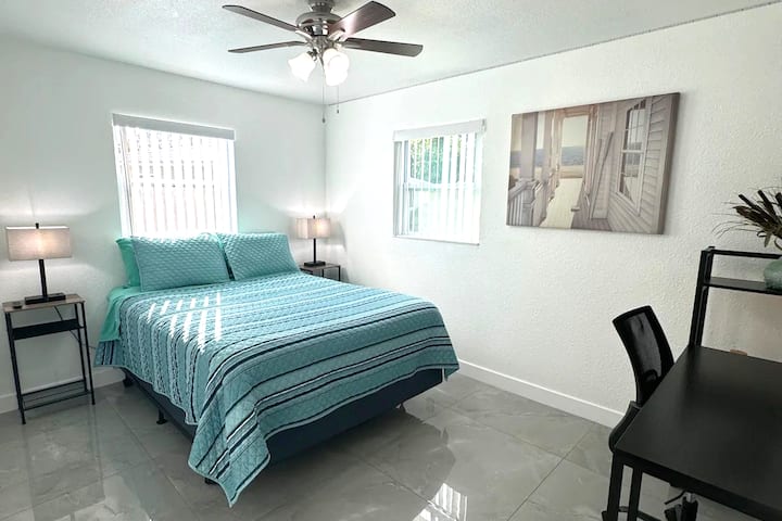 Large room with gray tile floor. Queen size bed with blue comforter. Across from bed is a black desk. There is a painting of a beach house on the right of the bed. 