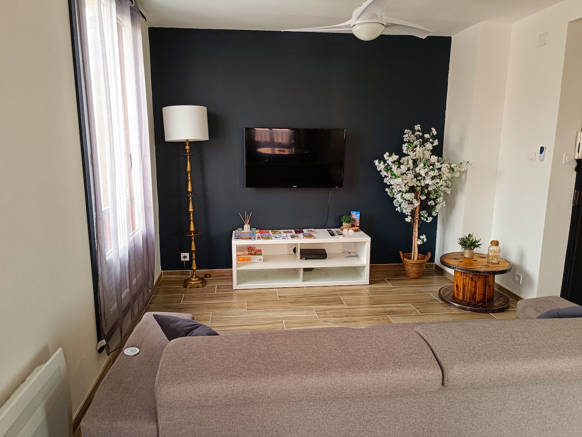 A living area features a neutral color palette, with a comfortable sofa facing a flat-screen television mounted on a minimalistic white media console. A tall lamp stands beside the window, and decorative artwork is displayed on a wooden coffee table. A faux plant adds a touch of greenery.