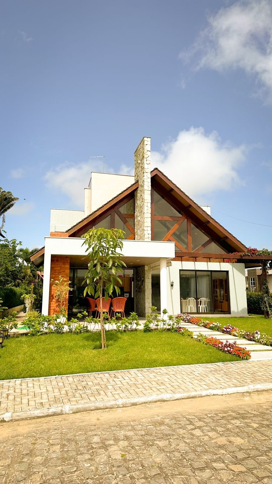 The exterior of the house showcases a modern design with a steeply pitched roof and large front windows. A well-manicured garden features colorful flowers and greenery. A welcoming entrance includes a covered porch with seating and stone accents, inviting guests to explore the property.