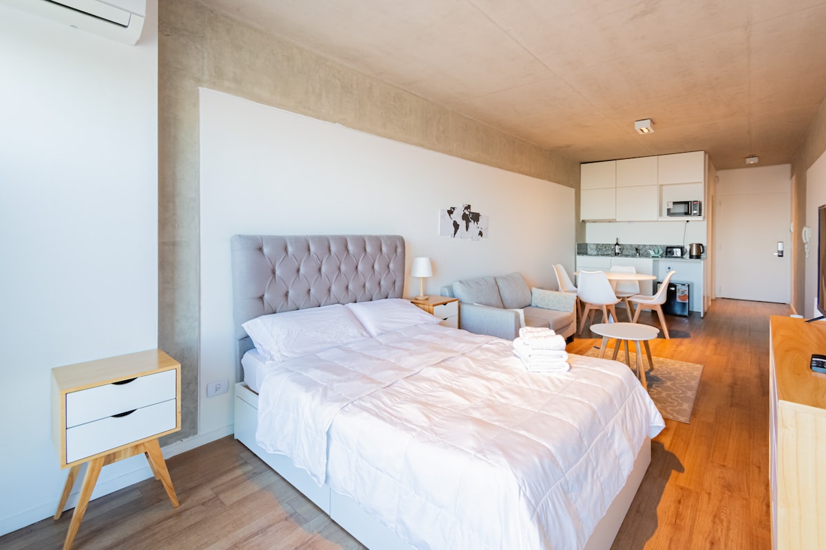 A modern studio apartment features a spacious bed with a soft headboard, complemented by a minimalist light wood nightstand. A comfortable sofa is positioned nearby, and a dining area with a table and four chairs is visible. Natural light illuminates the warm wood flooring.
